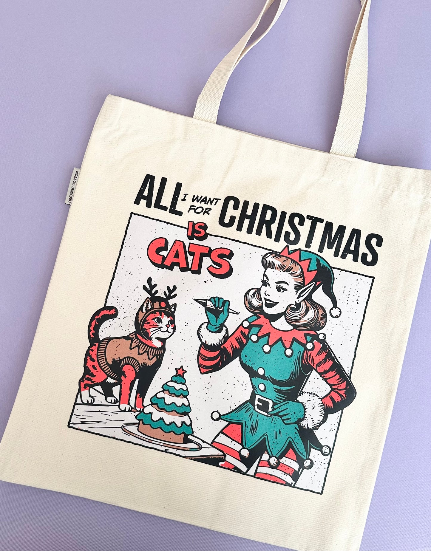 A product photo of a beige canvas tote bag with a vintage style illustration of a girl elf and a cat dressed as a reindeer decorating a Christmas tree cake. The text says All I Want For Christmas Is Cats