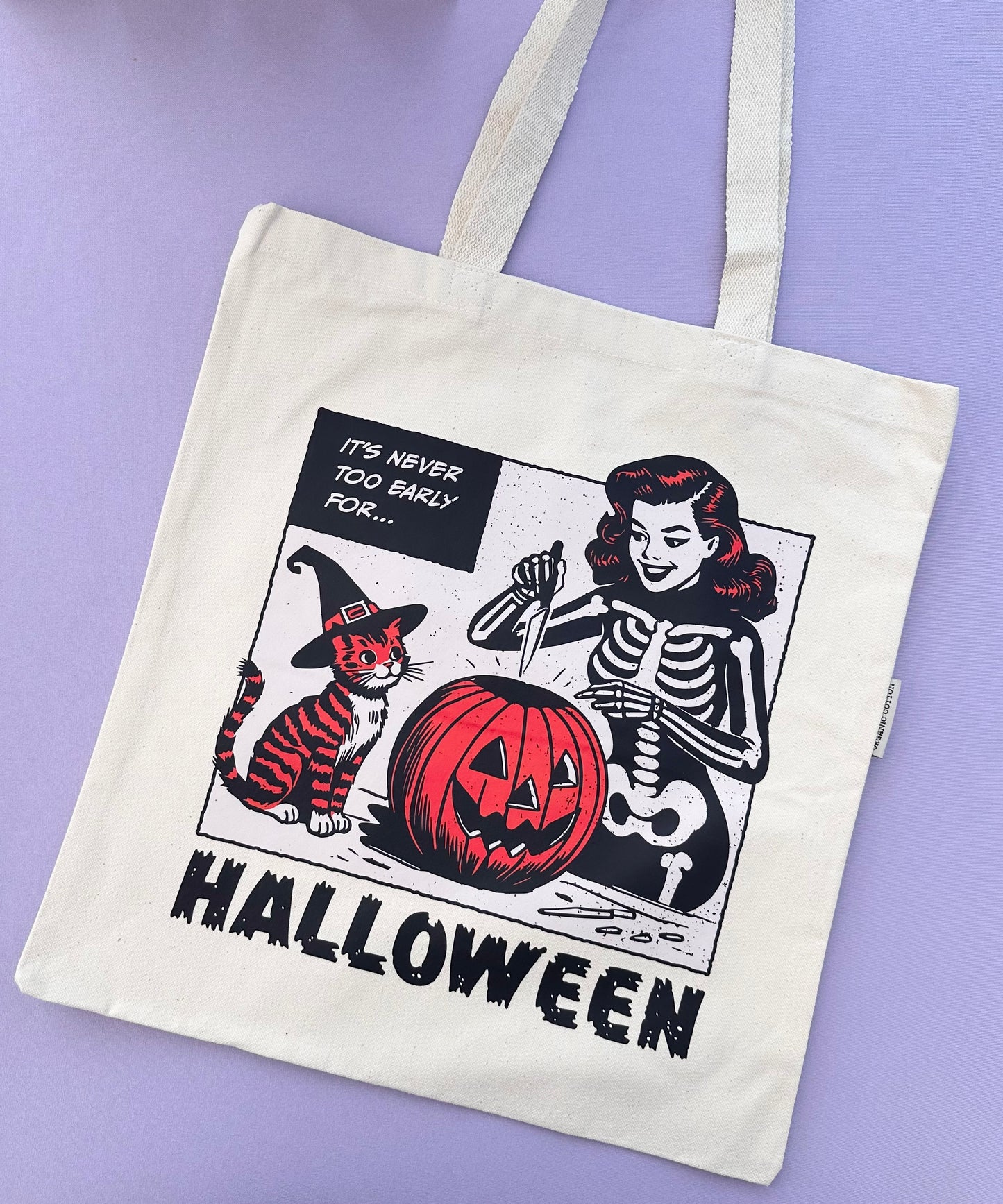 A product photo of a beige tote bag with a vintage style illustration of a girl dressed as a skeleton with an orange and black striped cat dressed as a witch. They are carving a jack-o-latern. The text says It's Never Too Early For Halloween