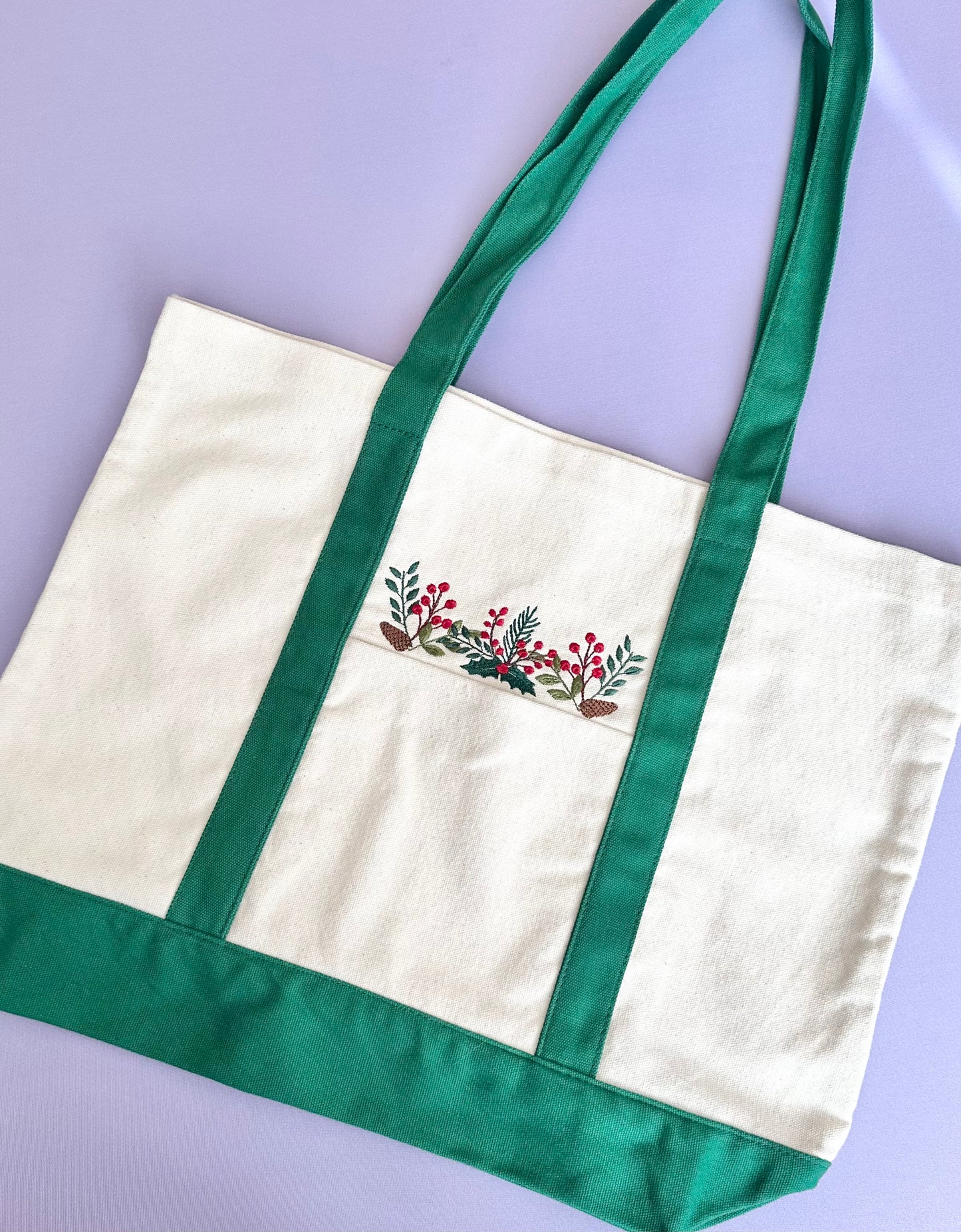 Product photo of a beige tote bag with green handles and bottom with an embroidered design of Poinsettias, pine cones, and hollies.