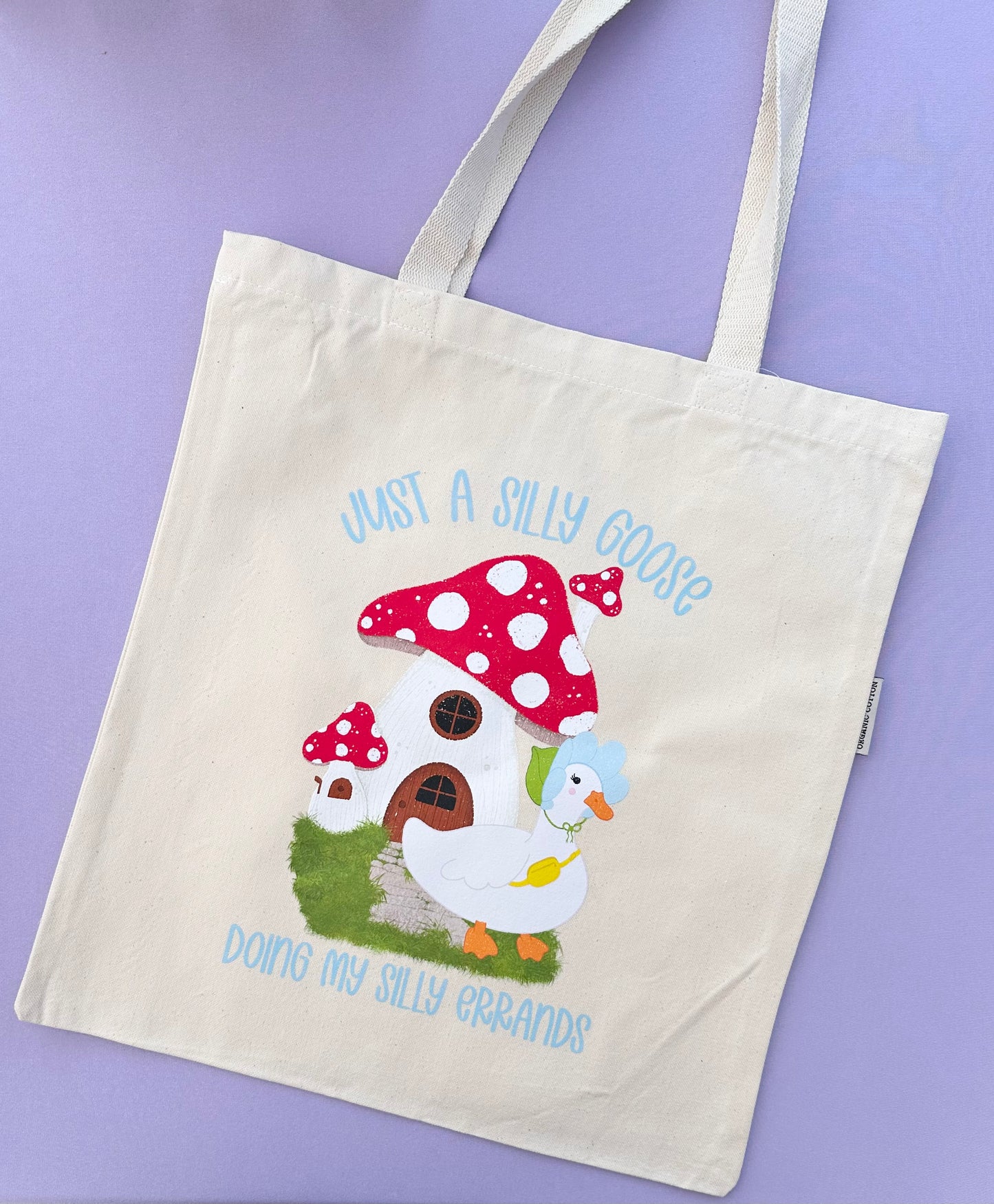 A product photo of a beige tote bag with an illustration of a mushroom house with a duck in front. The text says Just a Silly Goose Doing My Silly Errands.