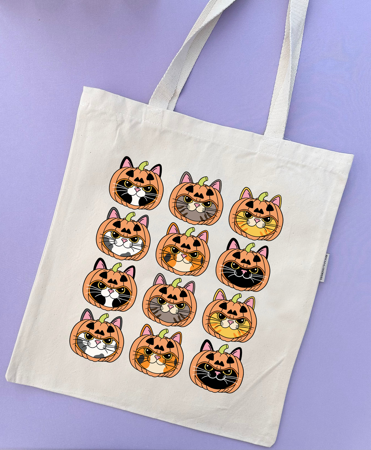 An off-white canvas tote bag with two shoulder straps, decorated with a grid of 12 cartoon pumpkin-and-cat faces in orange, black, white, gray, and yellow. The bag is photographed flat against a solid lavender-purple background.