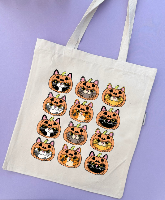 An off-white canvas tote bag with two shoulder straps, decorated with a grid of 12 cartoon pumpkin-and-cat faces in orange, black, white, gray, and yellow. The bag is photographed flat against a solid lavender-purple background.